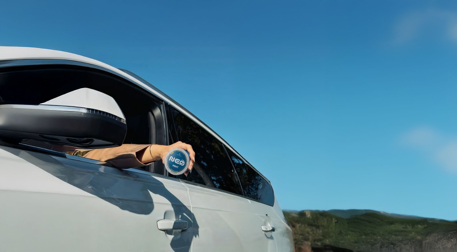 A hand holding a NEÖ tin out of a car window under a blue sky