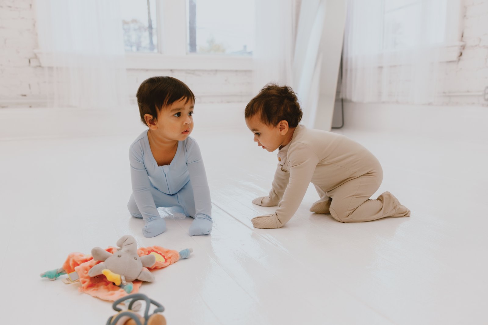 Two babies in crawl suits on a white floor